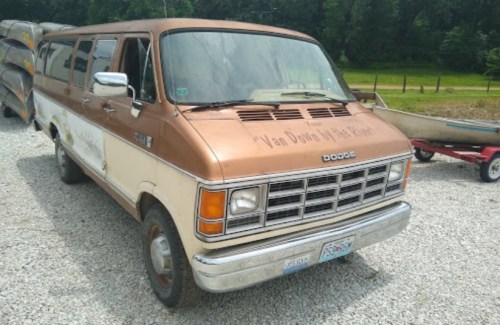 A vintage Dodge van with a brown and cream exterior parked on gravel, surrounded by canoes and greenery.