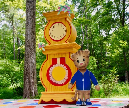 A colorful clock tower stands in a forest, with a cartoon tiger character in a blue jacket beside it.