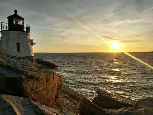 A lighthouse stands on rocky shore as the sun sets over the calm ocean, casting warm light across the scene.