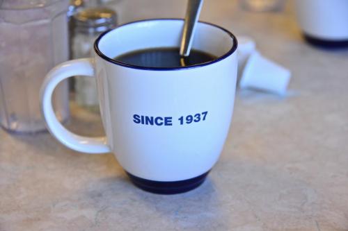 A white coffee mug with a blue rim, featuring the text "SINCE 1937," placed on a table with a glass of water nearby.