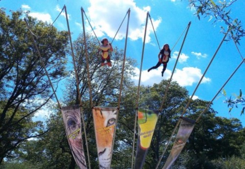 Two children are bouncing on a trampoline-like attraction against a blue sky with fluffy clouds and trees in the background.