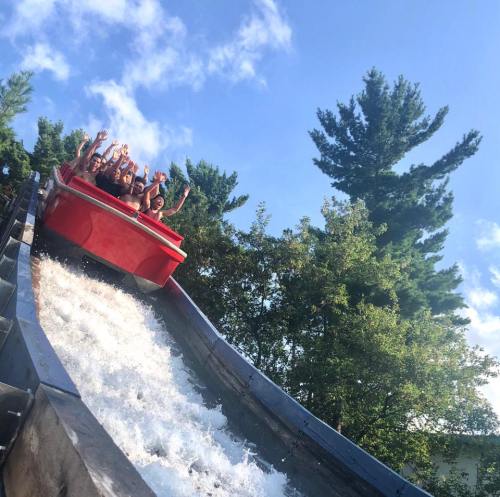 A group of people joyfully riding a water slide, splashing down amidst trees and a blue sky.