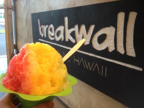 A colorful shaved ice dessert in a green cup, held in front of a sign that reads "breakwall" in Hawaii.