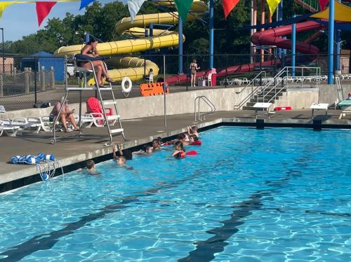 A lifeguard watches over swimmers at a pool with water slides in the background, colorful flags overhead.