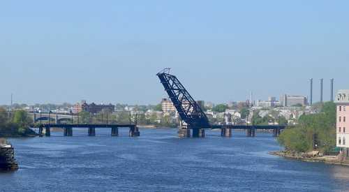 A raised drawbridge over a river, with city buildings and smokestacks in the background under a clear blue sky.