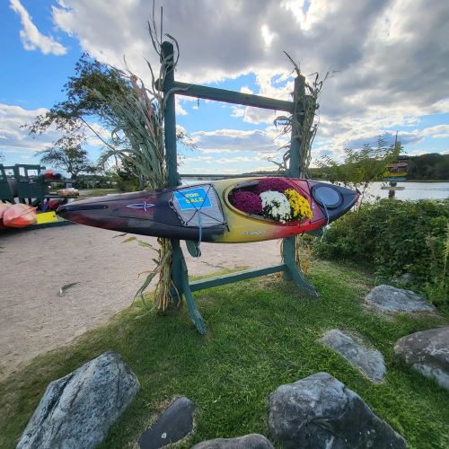 A colorful kayak adorned with flowers is displayed on a stand near a river, surrounded by greenery and rocks.