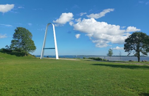 A white arch structure stands on a grassy area by a calm lake under a blue sky with fluffy clouds.