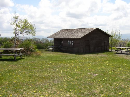 A small wooden cabin surrounded by green grass and picnic tables under a partly cloudy sky.