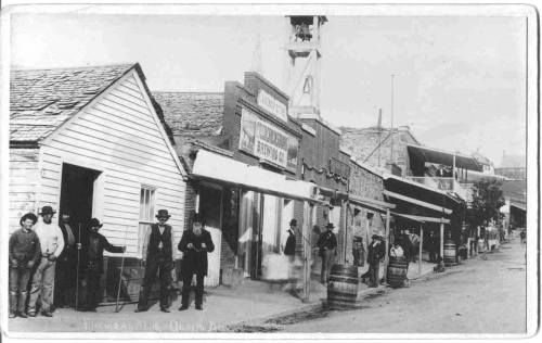 Historic black-and-white photo of a street scene with people and storefronts from the past.