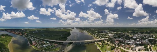 Aerial view of a river winding through a green landscape, with a bridge and a small town under a blue sky with clouds.