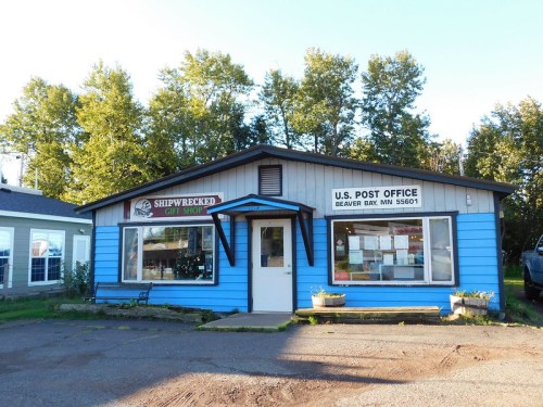 A blue building with a sign for "Shipwrecked Gift Shop" and a U.S. Post Office in Beaver Bay, MN, surrounded by trees.