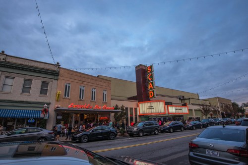 A street view of SCAD with a bustling crowd, cars parked, and a cloudy sky overhead. Lights adorn the buildings.