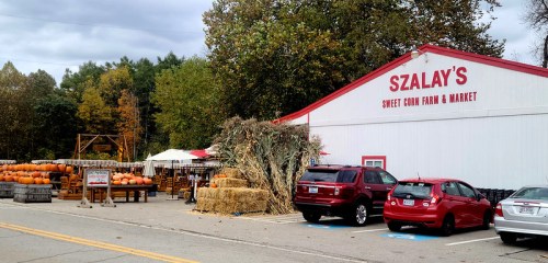 A farm market with pumpkins, hay bales, and cars parked outside a red and white building surrounded by trees.