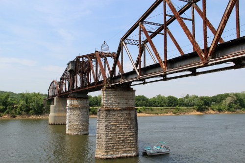 Rusty metal bridge spans a river, supported by stone pillars, with a small boat floating nearby under a clear blue sky.