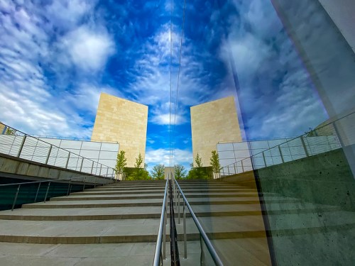 A staircase leads to two large stone structures, reflecting a blue sky with clouds in the glass surface.