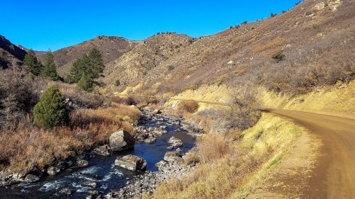 A winding dirt road alongside a stream, surrounded by hills and sparse vegetation under a clear blue sky.