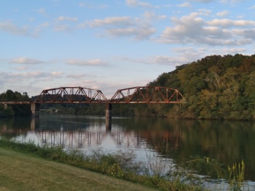 A rust-colored bridge spans a calm river, surrounded by lush greenery and a blue sky with scattered clouds.