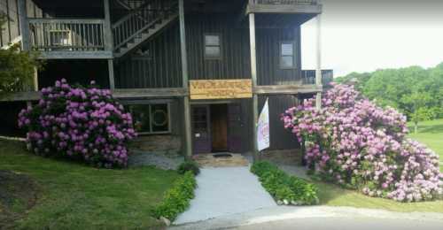 A rustic building with a wooden sign, surrounded by blooming pink rhododendrons and a welcoming pathway.