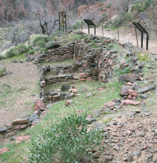 Ruins of stone structures surrounded by grass and rocky terrain, with informational signs nearby.