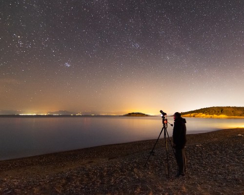 A person stands by the shore at night, using a telescope to observe a starry sky over calm waters.
