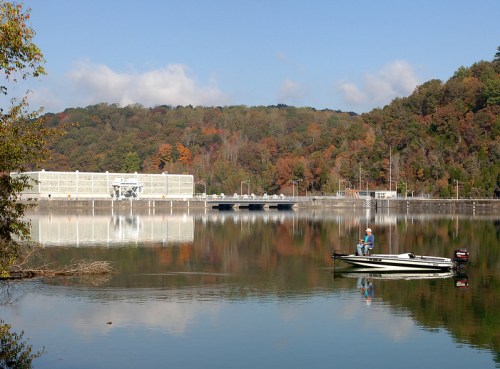 A serene lake scene with a boat and a person fishing, surrounded by autumn foliage and a dam in the background.