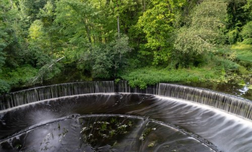 A serene waterfall cascades over a circular dam, surrounded by lush green trees and foliage.