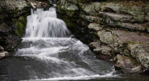 A serene waterfall cascading over rocks, surrounded by lush greenery and natural stone formations.