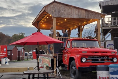 A vintage red truck parked near a wooden stage with lights, surrounded by small red buildings and a patio umbrella.