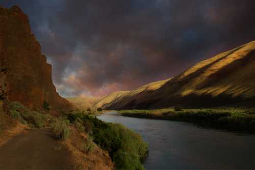 A serene river flows between rugged hills under a dramatic sky, with lush greenery lining the banks.