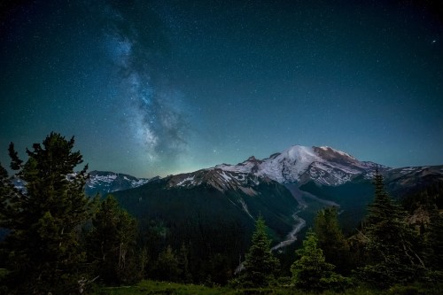 A stunning night sky filled with stars above a snow-capped mountain, surrounded by lush green trees.