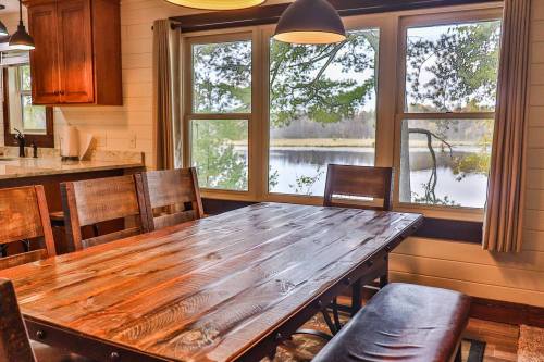 A rustic wooden dining table with chairs, overlooking a serene lake through large windows.