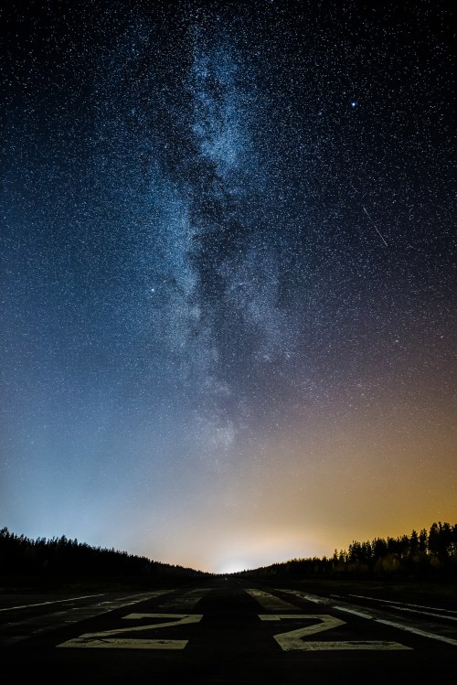 A starry night sky with the Milky Way above a dark runway, illuminated by a soft orange glow on the horizon.