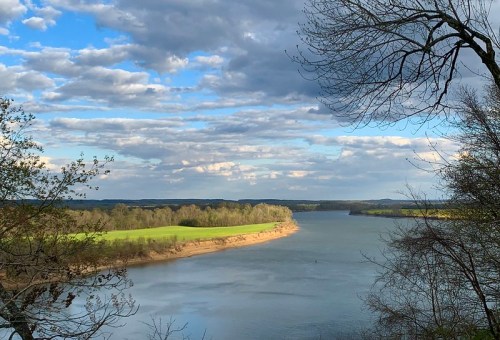 A serene river bends through lush green fields under a partly cloudy sky, framed by trees on the banks.