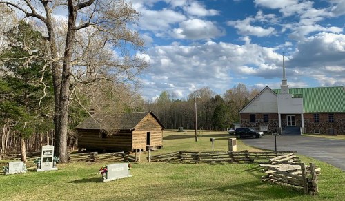A rural scene featuring a log cabin, a church with a steeple, gravestones, and a wooden fence under a cloudy sky.
