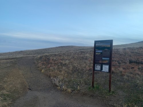 A trail sign stands beside a dirt path on a grassy hillside under a cloudy sky.