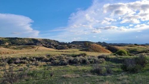 A scenic landscape featuring rolling hills, green grass, and a blue sky with scattered clouds.