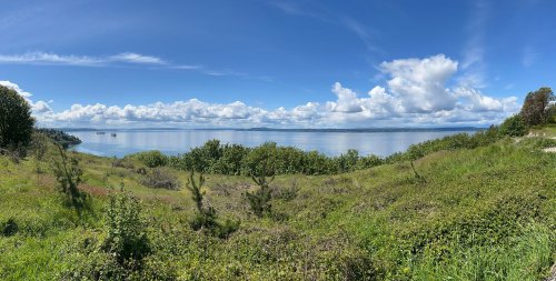 A panoramic view of a serene lake surrounded by lush greenery and a clear blue sky with scattered clouds.