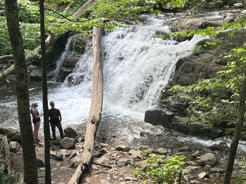 Two people stand by a waterfall surrounded by lush greenery and rocky terrain.