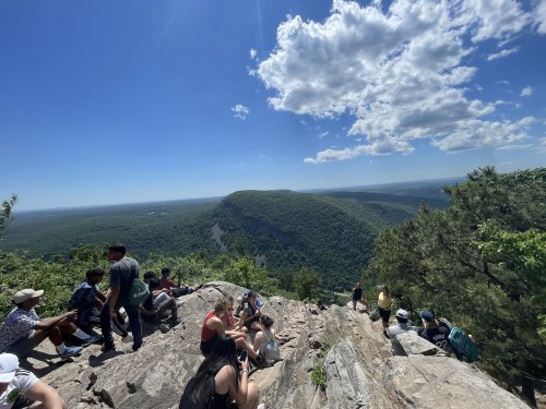 A group of people sitting on a rocky ledge, enjoying a scenic view of green mountains and a blue sky with clouds.