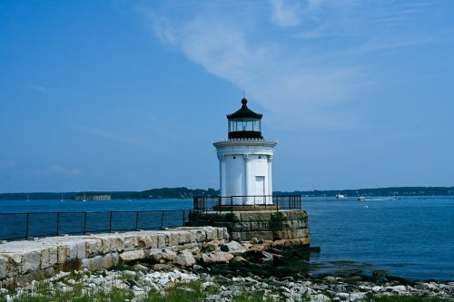 A white lighthouse stands on a rocky shore under a clear blue sky, with calm waters in the background.
