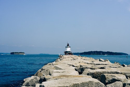A lighthouse stands at the end of a rocky pier, with calm blue waters and distant islands under a clear sky.