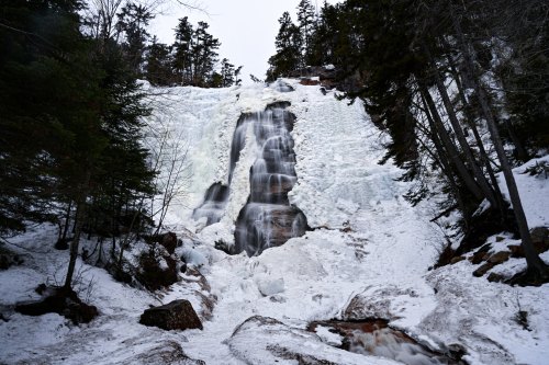 A frozen waterfall surrounded by snow-covered trees and rocky terrain, creating a serene winter landscape.