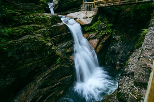 A serene waterfall cascades over rocky cliffs, surrounded by lush greenery and a wooden bridge above.