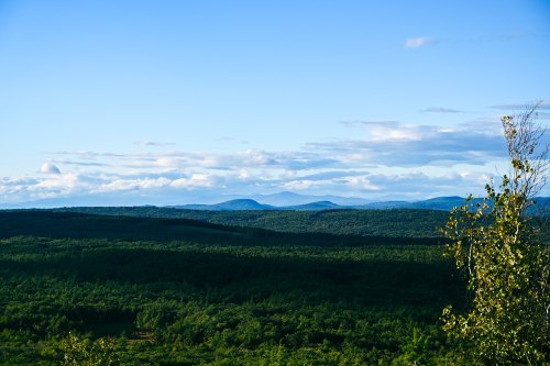 A panoramic view of rolling green hills and distant mountains under a clear blue sky with scattered clouds.