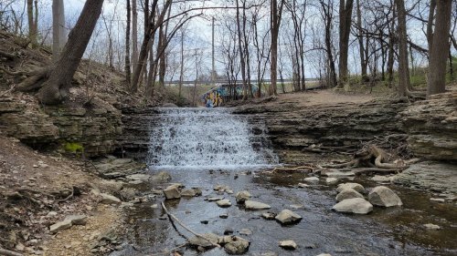 A small waterfall cascades over rocky terrain, surrounded by bare trees and a graffiti-covered wall in the background.