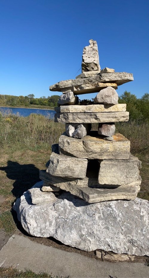 A stacked stone cairn beside a calm body of water, with clear blue skies and greenery in the background.