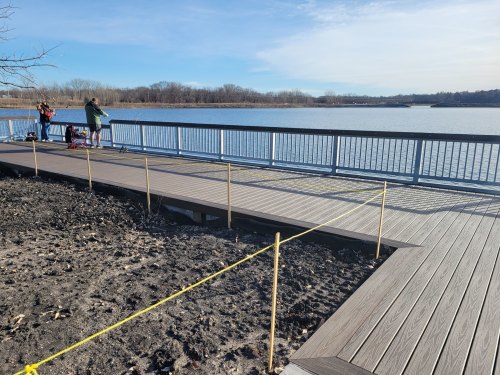 A wooden boardwalk extends over a calm lake, with people fishing in the distance and construction barriers nearby.