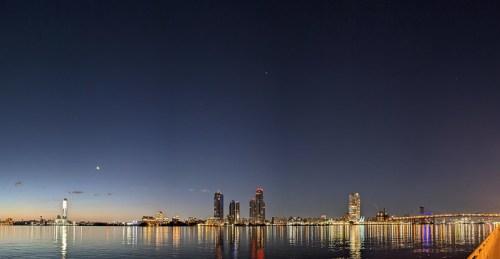 Panoramic view of a city skyline at night, reflecting on calm water with a crescent moon in the sky.