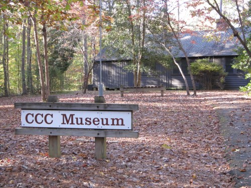 Sign for CCC Museum in a wooded area with fallen leaves and a dark building in the background.