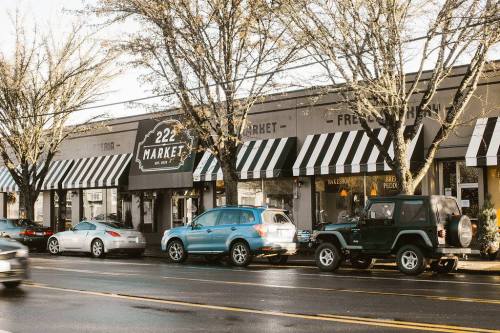 A street view of a market with striped awnings, parked cars, and trees lining the sidewalk on a rainy day.